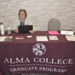A woman sits at a table for Alma College Graduate Programs, with brochures and a laptop on the table.