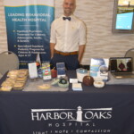 A person smiling behind a display table for Harbor Oaks Hospital, featuring brochures, small baskets, and treats. The backdrop highlights their behavioral health services.