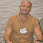 A person wearing a brown shirt and a name tag sits at a table with a laptop.