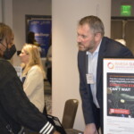 A man and a woman at an event discuss at a booth with a display about Invega Sustenna, a medication. The man is leaning on the table, and the woman is wearing a mask.