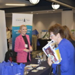 Person in a pink blazer stands at an informational booth with pamphlets and a banner. Another person in a blue coat looks at a brochure.