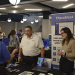 Three people at a business exhibition booth for TheraNest. A woman stands beside a display, a man in a white shirt talks to a visitor. Promotional materials scattered on the table.