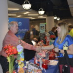 People are at a health fair, with a man in a red shirt shaking hands with a woman in a blue top across a table with brochures and decorations.