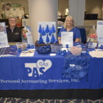 Two people sitting at a booth for Personal Accounting Services, Inc., featuring promotional items, brochures, and informational displays on a blue tablecloth.
