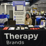 A person sits at a booth for TheraNest by Therapy Brands, featuring promotional materials and a banner that highlights their administrative software for therapy practices.