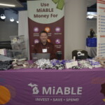 A man sits behind a table at a MiABLE booth with promotional items. A banner behind him lists financial benefits of MiABLE for housing, education, and more.
