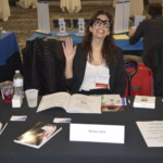 Person sitting at a table with books and papers, smiling and waving. The name tag reads "Bethany Bok.