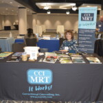 A woman sits at a table displaying materials for CCI MRT, with banners and brochures visible, in a conference setting.