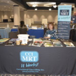 A woman sits at a table displaying materials for CCI MRT, with banners and brochures visible, in a conference setting.