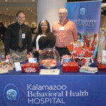 Three people stand behind a table showcasing giveaways and pamphlets at a Kalamazoo Behavioral Health Hospital booth.