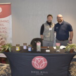 Two men stand behind a table with marketing materials and plants. A banner reads "Reclaiming Lives, Healing Families." The table features the Rose Hill Center logo.