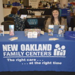 A representative sits behind a table for New Oakland Family Centers, featuring brochures, business cards, and branded items.