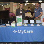 Two people stand behind a table displaying MyCare products at an event. The table has several devices and informational signs.