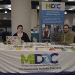 Two people sit at an informational table for the Michigan Developmental Disabilities Council, featuring brochures and a banner reading "Our Community Includes Everyone.