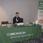 A person sits behind a booth table covered with a green tablecloth labeled "Bronson Behavioral Health Hospital." The display includes brochures, mugs, and a sign about an inpatient older adult unit.