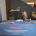 A woman sits at a booth for "mend," promoting mental and behavioral healthcare services. The table displays brochures, a phone, a water bottle, and a backdrop features hangers and chairs.