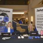 Two women at a Rehmann booth, seated at a table with promotional materials and laptops. Banners display company services like accounting and human resource solutions.