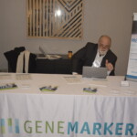 Man seated at a table with a "GeneMarkers" tablecloth, using a laptop and giving a thumbs-up. Various informational materials are spread across the table.