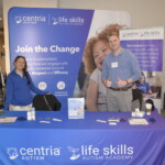 Two people stand behind a table with Centria Autism and Life Skills Academy banners, displaying promotional materials. The tablecloth features the logos of both organizations.