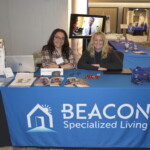 Two people sit at a table with brochures and items, promoting Beacon Specialized Living. The table is covered with a blue tablecloth featuring the company logo.