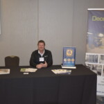 A man sits at a booth with brochures on the table and promotional banners behind him for Decofloor and NORIX.