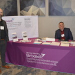 Two men stand at a Brixadi booth with brochures and promotional materials. A pink tablecloth displays information about the medication.