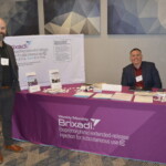 Two men stand at a Brixadi booth with brochures and promotional materials. A pink tablecloth displays information about the medication.