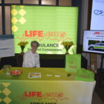 A person sits at a booth for LIFE EMS Ambulance at an event. The table has brochures and a display behind reads "Service and Compassion". A monitor shows information about health services.