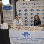 Two representatives sit at a Heartland Center for Autism booth with promotional materials on a table. A banner and a standee display information about their autism treatment programs.