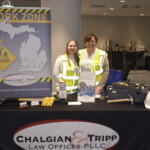Two people in safety vests stand behind a table with a Chalgian & Tripp banner, hard hats, and brochures. The setting appears to be a conference or event space.