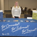 Person standing behind a table covered with a blue cloth. The cloth displays the words "Get Connected! Get Informed! Get Trained!" The table has brochures and pens.