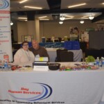 ???????????????????????????????????? Two people sitting at a booth for Bay Human Services, with brochures, candy, and water bottles on the table. Banners and other booths are visible in the background.