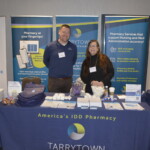 Two people stand behind an exhibit table with promotional items. Banners display "Tarrytown" and pharmacy services related text.
